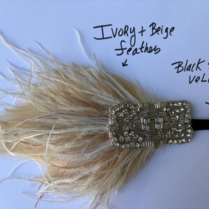 May include: A headpiece featuring ivory and beige feathers attached to a rectangular, silver-toned embellishment covered in clear rhinestones and beads. A black stretch velvet band extends from the side. Text reads: Ivory + Beige feathers and Black stretch velvet.