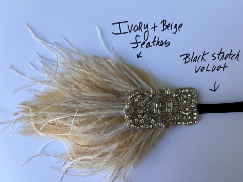 May include: A headpiece featuring ivory and beige feathers attached to a rectangular, silver-toned embellishment covered in clear rhinestones and beads. A black stretch velvet band extends from the side. Text reads: Ivory + Beige feathers and Black stretch velvet.