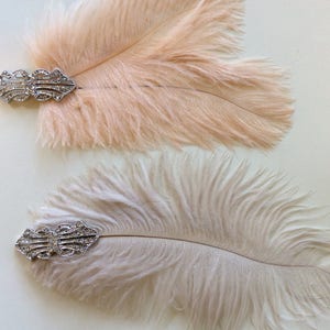 May include: Two decorative hair clips featuring soft, fluffy ostrich feathers in pale peach and cream tones. Each feather is attached to a silver-toned metal base adorned with clear, sparkling rhinestones in an intricate, symmetrical design.