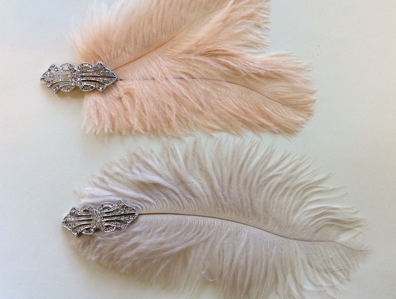 May include: Two decorative hair clips featuring soft, fluffy ostrich feathers in pale peach and cream tones. Each feather is attached to a silver-toned metal base adorned with clear, sparkling rhinestones in an intricate, symmetrical design.