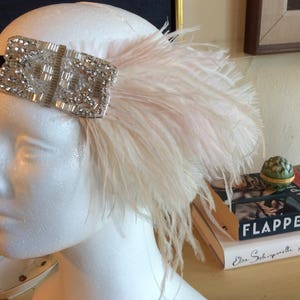 May include: A headpiece featuring a rectangular silver-toned embellishment covered in clear crystals and beads, attached to a black band. A large, soft, pale pink ostrich feather plume extends from the side of the crystal-encrusted ornament.