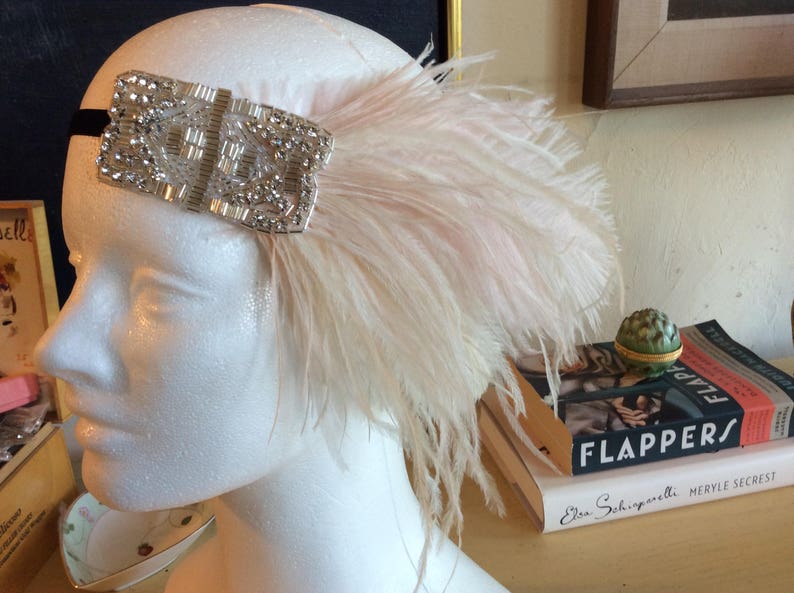 May include: A headpiece featuring a rectangular silver-toned embellishment covered in clear crystals and beads, attached to a black band. A large, soft, pale pink ostrich feather plume extends from the side of the crystal-encrusted ornament.