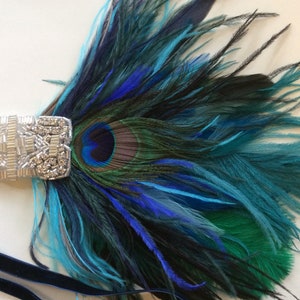 May include: A decorative hair accessory featuring a silver-toned rectangular bar embellished with clear crystals and beads. Attached to the bar is a cluster of vibrant peacock feathers in shades of deep blue, teal, and green, secured by a dark blue velvet ribbon.