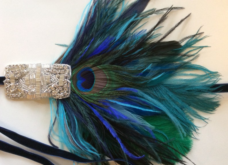 May include: A decorative hair accessory featuring a silver-toned rectangular bar embellished with clear crystals and beads. Attached to the bar is a cluster of vibrant peacock feathers in shades of deep blue, teal, and green, secured by a dark blue velvet ribbon.