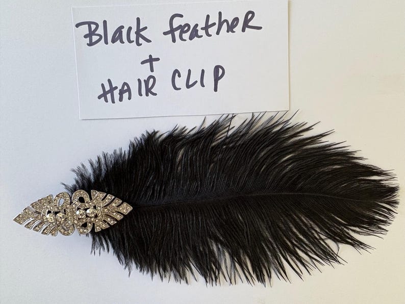 May include: A black feather hair clip featuring a silver-toned metal accent piece adorned with clear rhinestones. A white rectangular card is positioned above the item with the handwritten text Black feather + HAIR CLIP in black ink.
