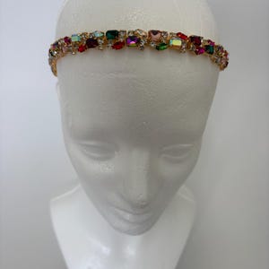 May include: A thin gold-tone headband featuring a continuous row of multi-colored rectangular and round crystals. The embellishments include shades of red, green, pink, and iridescent stones set against a metallic gold frame that encircles the head.