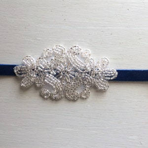 Peut inclure: Un collier ras du cou en velours bleu marine avec un motif floral en perles argent&eacute;es. La d&eacute;coration perl&eacute;e est centr&eacute;e sur le collier et est compos&eacute;e de petites perles brillantes. Le collier est pr&eacute;sent&eacute; sur une surface en bois blanc.