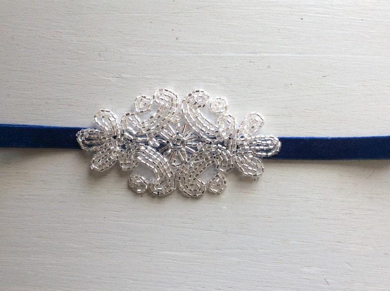Peut inclure: Un collier ras du cou en velours bleu marine avec un motif floral en perles argent&eacute;es. La d&eacute;coration perl&eacute;e est centr&eacute;e sur le collier et est compos&eacute;e de petites perles brillantes. Le collier est pr&eacute;sent&eacute; sur une surface en bois blanc.