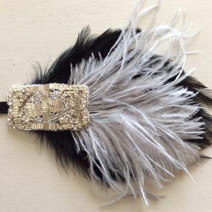 May include: A black and silver feather hair accessory featuring a rectangular embellishment covered in clear rhinestones and silver beads. The piece is attached to a thin black elastic band and displays layered black and light grey feathers.
