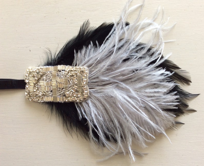 May include: A black and silver feather hair accessory featuring a rectangular embellishment covered in clear rhinestones and silver beads. The piece is attached to a thin black elastic band and displays layered black and light grey feathers.