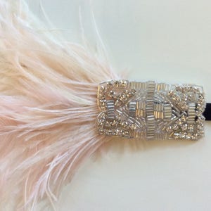 May include: A headband featuring a rectangular silver-toned embellishment decorated with clear crystals and glass beads. A cluster of soft, pale pink ostrich feathers is attached to the side of the beaded piece, which is secured to a thin black band.