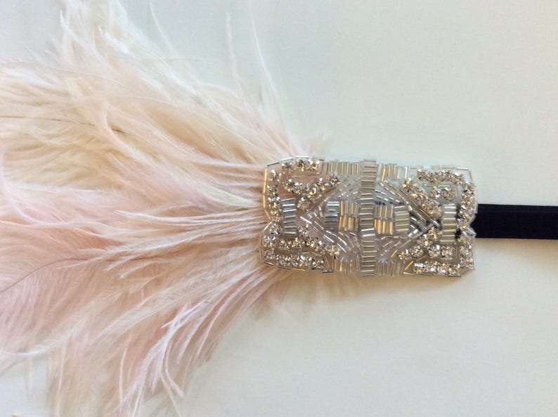 May include: A headband featuring a rectangular silver-toned embellishment decorated with clear crystals and glass beads. A cluster of soft, pale pink ostrich feathers is attached to the side of the beaded piece, which is secured to a thin black band.