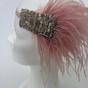 May include: A pale pink headband featuring a rectangular embellishment made of clear crystals and silver-toned beads. The accessory is accented with soft, wispy pale pink feathers that extend outwards from the side of the beaded centrepiece.