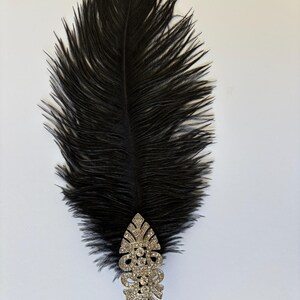 May include: A long, black ostrich feather plume attached to a silver-toned, elongated decorative brooch. The brooch features a symmetrical, intricate metalwork design embedded with numerous small, clear, sparkling rhinestones.