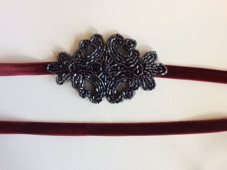 Peut inclure: Un bandeau en velours bordeaux orn&eacute; d'une d&eacute;coration florale en perles gris fonc&eacute;. La d&eacute;coration perl&eacute;e est centr&eacute;e sur le bandeau, cr&eacute;ant un accent d&eacute;coratif. Un accessoire simple et &eacute;l&eacute;gant.