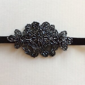 Peut inclure: Un collier ras du cou noir orn&eacute; de perles. Le collier est compos&eacute; d'un ruban de velours avec un motif floral fait de petites perles gris fonc&eacute;. L'ornement de perles est centr&eacute; sur le ruban.