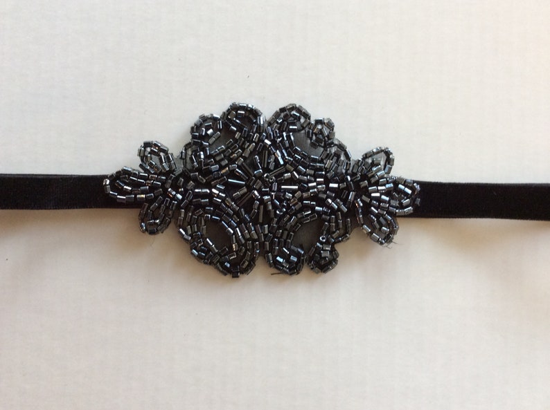 Peut inclure: Un collier ras du cou noir orn&eacute; de perles. Le collier est compos&eacute; d'un ruban de velours avec un motif floral fait de petites perles gris fonc&eacute;. L'ornement de perles est centr&eacute; sur le ruban.