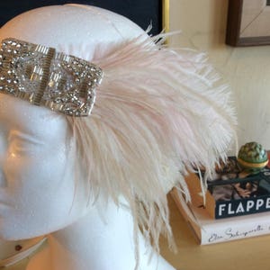May include: A headpiece featuring a rectangular silver-toned embellishment covered in clear crystals and beads, attached to a black elastic band. A large, soft, pale pink ostrich feather plume extends from the side of the crystal ornament.