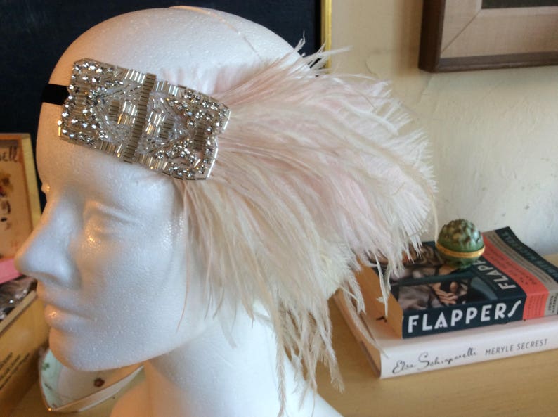 May include: A headpiece featuring a rectangular silver-toned embellishment covered in clear crystals and beads, attached to a black elastic band. A large, soft, pale pink ostrich feather plume extends from the side of the crystal ornament.