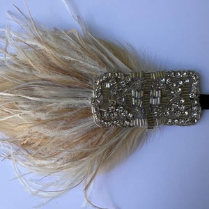 May include: A decorative headband featuring a cluster of light beige ostrich feathers attached to a rectangular silver-toned embellishment. The rectangular piece is densely covered in clear faceted crystals and metallic silver beads.