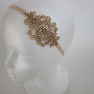 Peut inclure: Un bandeau dor&eacute; orn&eacute; de perles avec un motif floral. Le bandeau est fait d'un tissu beige doux et pr&eacute;sente un groupe de perles dor&eacute;es en forme de motif floral sur le c&ocirc;t&eacute;. Le bandeau est pr&eacute;sent&eacute; sur une t&ecirc;te de mannequin blanche.