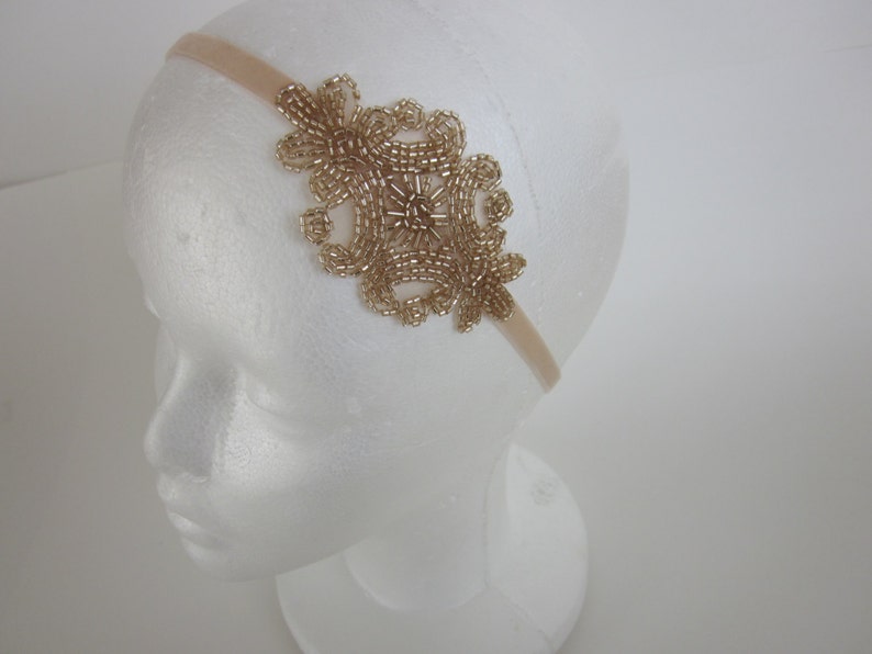 Peut inclure: Un bandeau dor&eacute; orn&eacute; de perles avec un motif floral. Le bandeau est fait d'un tissu beige doux et pr&eacute;sente un groupe de perles dor&eacute;es en forme de motif floral sur le c&ocirc;t&eacute;. Le bandeau est pr&eacute;sent&eacute; sur une t&ecirc;te de mannequin blanche.