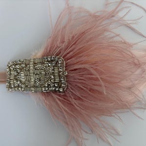 May include: A dusty pink feather headband featuring a rectangular silver-toned embellishment. The central piece is decorated with clear faceted crystals and metallic silver beads arranged in a geometric pattern, attached to a matching pink elastic band.