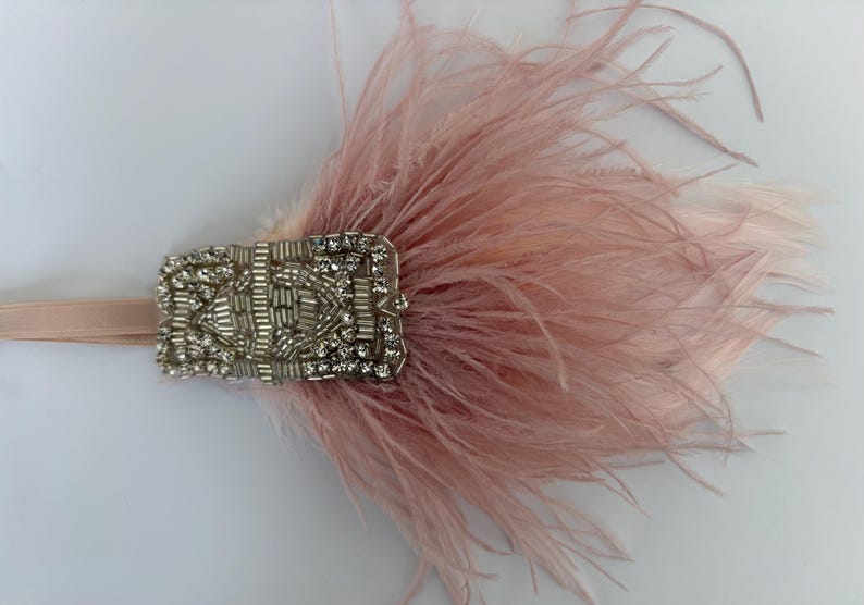 May include: A dusty pink feather headband featuring a rectangular silver-toned embellishment. The central piece is decorated with clear faceted crystals and metallic silver beads arranged in a geometric pattern, attached to a matching pink elastic band.