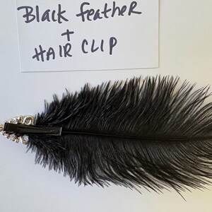 May include: A black feather hair clip featuring a long, soft black feather attached to a silver-toned metal base with clear crystal accents. A white card above the accessory displays the handwritten text: Black feather + HAIR CLIP.
