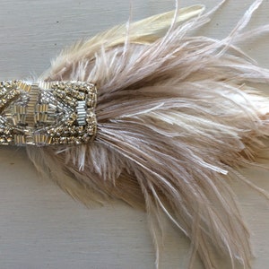May include: A headband featuring a rectangular embellishment adorned with clear crystals and silver metallic beads, paired with a cluster of soft, light beige ostrich feathers attached to a thin, matching elastic band.