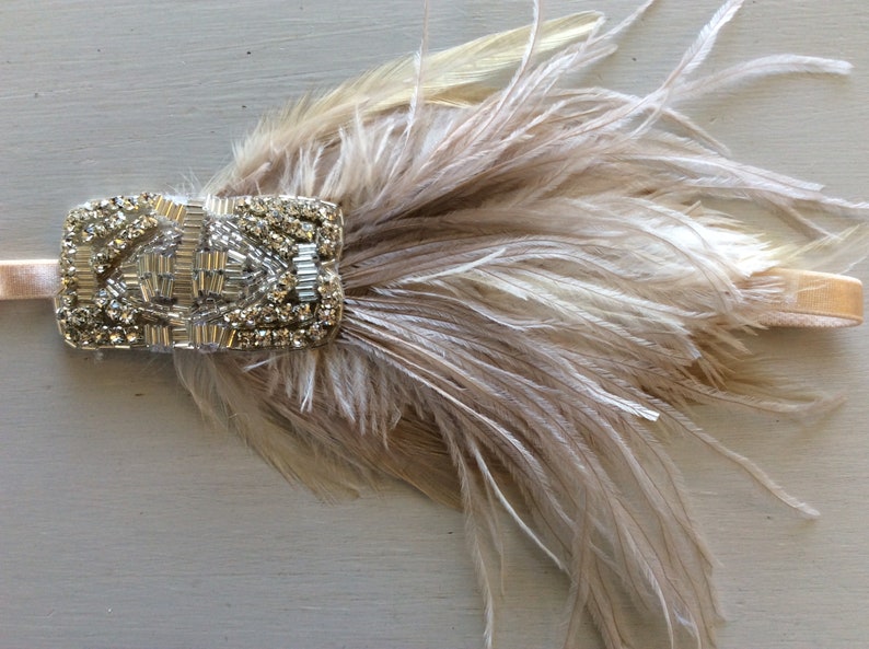 May include: A headband featuring a rectangular embellishment adorned with clear crystals and silver metallic beads, paired with a cluster of soft, light beige ostrich feathers attached to a thin, matching elastic band.