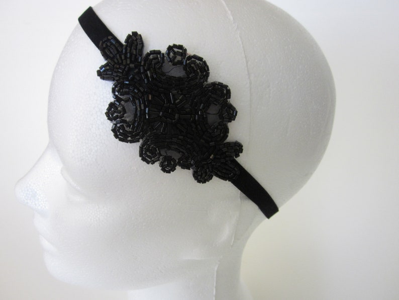 Peut inclure: Bandeau noir orn&eacute; de perles. Il pr&eacute;sente un grand motif floral sur le c&ocirc;t&eacute;, compos&eacute; de petites perles sombres, cr&eacute;ant un aspect textur&eacute; et d&eacute;taill&eacute;. Le bandeau est pr&eacute;sent&eacute; sur une t&ecirc;te de mannequin blanche.