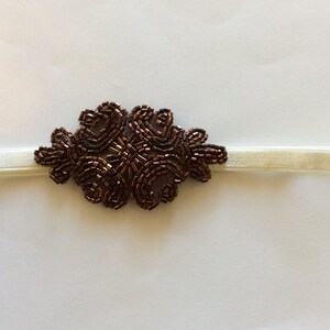 Peut inclure: Un bandeau &eacute;lastique ivoire avec un motif floral central. L'ornement floral est compos&eacute; de petites perles marron fonc&eacute;, cr&eacute;ant un &eacute;l&eacute;ment d&eacute;coratif textur&eacute;. Le bandeau est adapt&eacute; &agrave; diverses occasions.