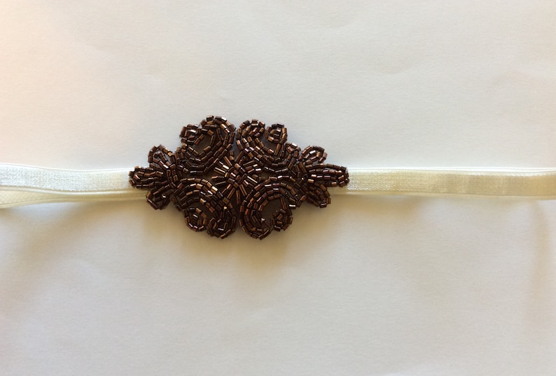 Peut inclure: Un bandeau &eacute;lastique ivoire avec un motif floral central. L'ornement floral est compos&eacute; de petites perles marron fonc&eacute;, cr&eacute;ant un &eacute;l&eacute;ment d&eacute;coratif textur&eacute;. Le bandeau est adapt&eacute; &agrave; diverses occasions.