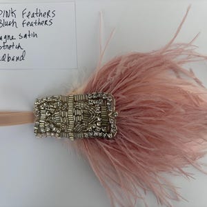 May include: A stretch headband featuring a rectangular embellishment with silver beads and clear crystals, accented by soft pink and blush feathers. A white card is present with the handwritten text: ALL PINK FEATHERS ALL BLUSH FEATHERS + Champagne satin stretch headband.
