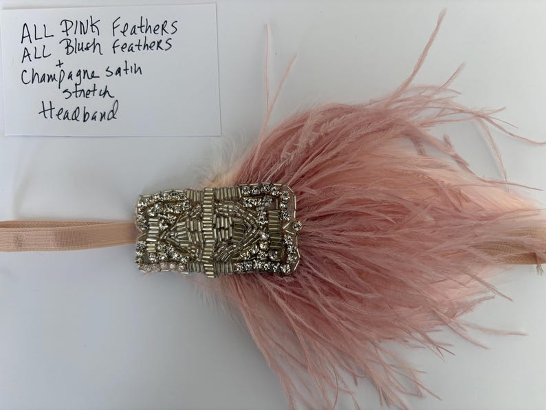 May include: A stretch headband featuring a rectangular embellishment with silver beads and clear crystals, accented by soft pink and blush feathers. A white card is present with the handwritten text: ALL PINK FEATHERS ALL BLUSH FEATHERS + Champagne satin stretch headband.