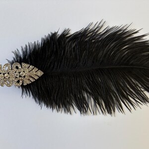 May include: A decorative hair accessory featuring a long, black ostrich feather attached to a silver-toned metal clip. The clip is shaped like an elongated leaf or marquise design and is encrusted with numerous small, clear, sparkling rhinestones.