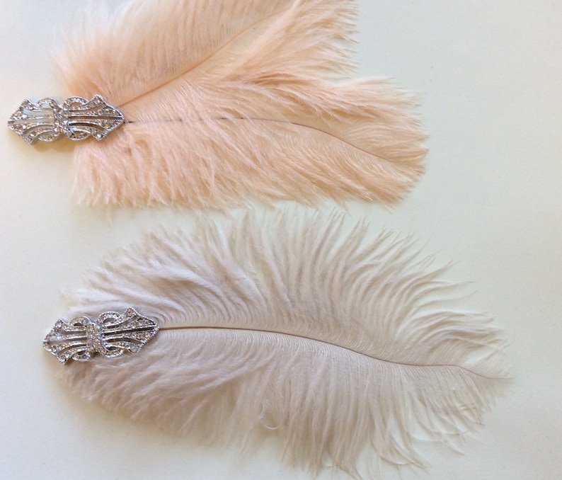 May include: Two decorative hair clips featuring soft, fluffy feathers in pale peach and light beige. Each feather is attached to a silver-toned metal base adorned with clear, sparkling rhinestones arranged in a symmetrical, geometric pattern.