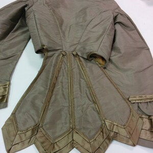 May include: An olive-green vintage jacket with long sleeves and a distinctive flared skirt. The jacket has satin trim and a high collar. The back view highlights the detailed tailoring and decorative features, providing a look at historical fashion.