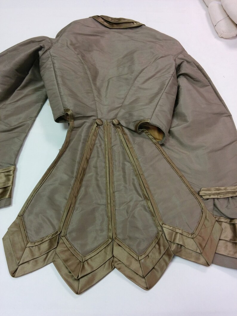 May include: An olive-green vintage jacket with long sleeves and a distinctive flared skirt. The jacket has satin trim and a high collar. The back view highlights the detailed tailoring and decorative features, providing a look at historical fashion.