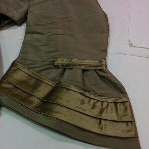 May include: Olive green fabric with a layered design. The fabric has a sheen and appears to be silk or a similar material. The bottom edge features multiple horizontal bands of a darker olive colour, creating a textured effect. The garment appears to be part of a dress or top.