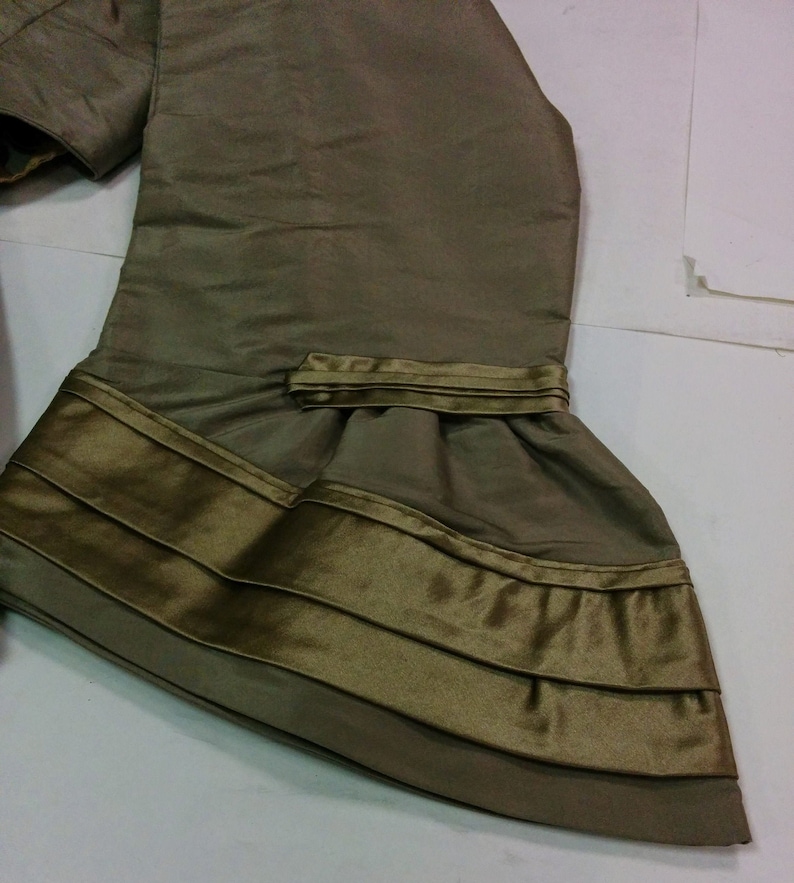 May include: Olive green fabric with a layered design. The fabric has a sheen and appears to be silk or a similar material. The bottom edge features multiple horizontal bands of a darker olive colour, creating a textured effect. The garment appears to be part of a dress or top.
