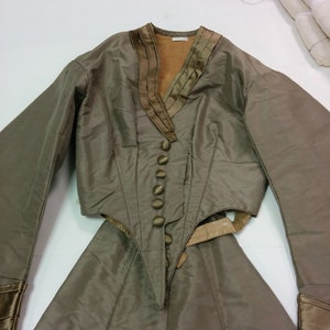 May include: An olive green two-piece outfit. The jacket features a V-neck with decorative trim, long sleeves, and a buttoned front. The skirt is a matching olive green colour.