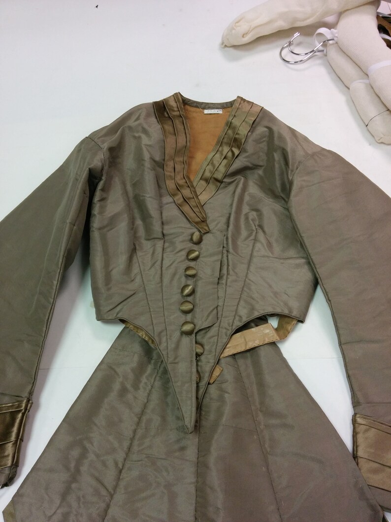 May include: An olive green two-piece outfit. The jacket features a V-neck with decorative trim, long sleeves, and a buttoned front. The skirt is a matching olive green colour.