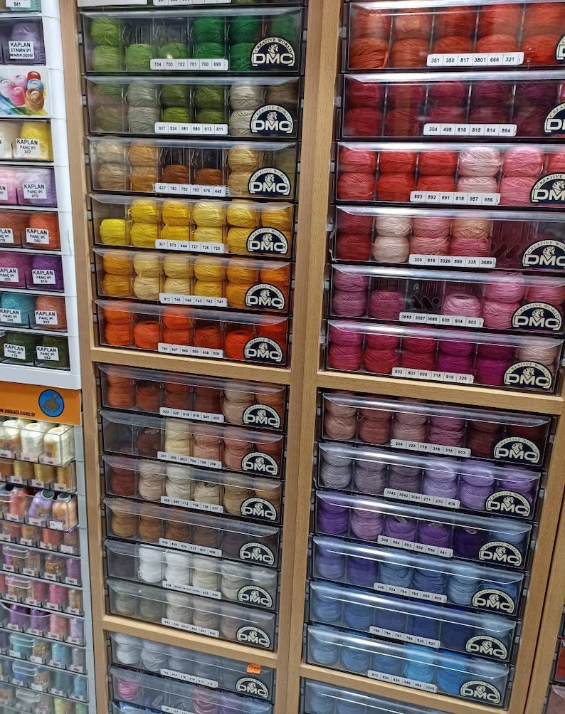 May include: A wall of drawers filled with colorful spools of DMC embroidery floss. Each drawer is labeled with a DMC color number.