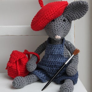 May include: A grey crocheted mouse wearing a red beret and blue dungarees, holding a paintbrush. The mouse is sitting on a white surface and has a red crocheted bag next to it.