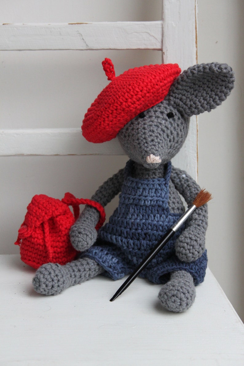 May include: A grey crocheted mouse wearing a red beret and blue dungarees, holding a paintbrush. The mouse is sitting on a white surface and has a red crocheted bag next to it.