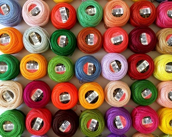 May include: A collection of colourful embroidery floss spools with DMC branding. The spools are arranged in a grid pattern on a white surface.