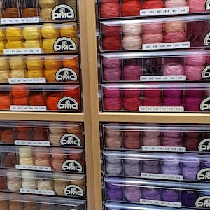 May include: A close-up view of a wall of drawers filled with spools of embroidery floss. The spools are organized by color and numbered. The DMC logo is visible on the drawers.