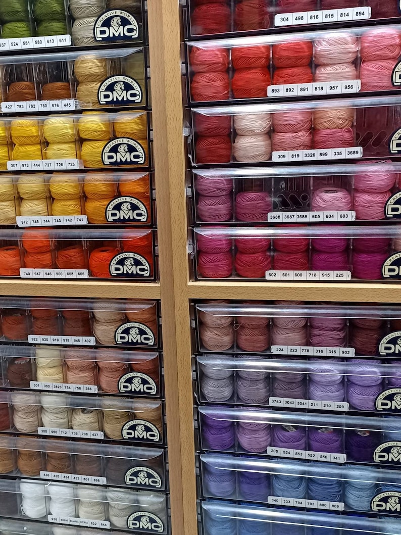 May include: A close-up view of a wall of drawers filled with spools of embroidery floss. The spools are organized by color and numbered. The DMC logo is visible on the drawers.